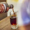 A dark liquid is being poured from a bottle into a glass. Produktbild-Vorschau