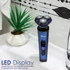 A blue electric shaver with a digital display sits on a white sink next to a potted plant. The background features a tiled wall with a decorative pattern. Produktbild-Vorschau