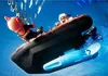 A diver in an orange helmet and black suit is being pulled into a black and orange inflatable boat by another person. A blue torpedo-like object is attached to the underside of the boat. Náhľad obrázku produktu
