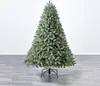 A green artificial Christmas tree with a flocked appearance, standing on a black metal stand on a gray wooden floor against a white wall. Produktbild-Vorschau