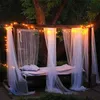 A bed with sheer curtains and string lights set up outdoors under a tree at dusk. Produktbild-Vorschau