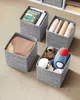 Four fabric storage boxes are arranged on a light-colored floor. One box contains books, another contains rolled towels, a third contains a folded blanket, and the fourth contains stuffed toys. Produktbild-Vorschau