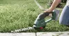 A person is using a cordless hedge trimmer to trim grass along a paved path. Produktbild-Vorschau