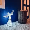 A lit angel decoration stands in the snow next to a basket. Náhled obrázku produktu