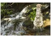 A wooden mechanical model of a hurdy-gurdy is placed on rocks next to a flowing stream in a natural setting. Produktbild-Vorschau