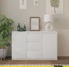 A white sideboard with three drawers and two doors, decorated with a framed picture, two green bottles, a green vase, and a white lamp. A large green plant in a patterned pot is on the left side of the sideboard. Náhled obrázku produktu