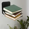 A black metal shelf holding a stack of four books against a light gray wall. A plant is visible in the lower right corner. Produktbild-Vorschau