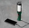 A green and black LED work light is shown charging a black smartphone on a gray concrete surface. Produktbild-Vorschau
