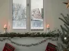 A festive Christmas mantelpiece decorated with a garland, candles, and stockings, with a window showing a snowy winter scene. Produktbild-Vorschau