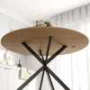 The underside of a round wooden dining table with a black metal base, shown in a kitchen setting with white cabinets and a window with sheer curtains in the background. Produktbild-Vorschau