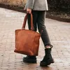 A person walking on a brick path, holding a brown leather tote bag. Voir les images du produit