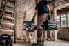 A person in work clothes is holding a Metabo tool case and a miter saw in a construction site. Podgląd zdjęcia produktu