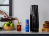 A hand is pouring a dark beverage into a glass filled with ice. Next to the glass are a bottle of Pepsi syrup and a SodaStream machine. A basket of green apples is also visible. Produktbild-Vorschau