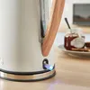 A close-up of a white kettle with a wooden handle, sitting on a wooden surface next to a jar of jam and toast. Podgląd zdjęcia produktu