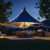 An outdoor patio scene at night with a large blue tent-like structure, a dining table set for a meal, and a grill. Colorful string lights are draped around the structure. Produktbild-Vorschau