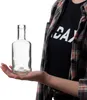 A person's hand is holding a clear glass bottle. Voir les images du produit