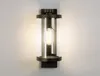 A modern outdoor wall light with a clear glass cylinder and a dark metal frame, mounted on a white wall. Produktbild-Vorschau