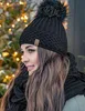 A woman wearing a black knitted hat with a pom-pom and a black scarf, with a blurred background of Christmas lights. Voir les images du produit