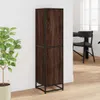 A tall, narrow cabinet with two doors, made of wood-look material with a black metal frame, stands against a white wall next to a potted plant. In the background, a dining table and chairs are visible through a window. Podgląd zdjęcia produktu