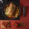A cooked roast with vegetables and a detachable handle for cookware, arranged on a stovetop and a red surface. Anteprima dell’immagine prodotto