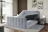 A modern bedroom with a grey upholstered bed frame that has its storage compartment open, revealing the mattress base and hydraulic lift mechanism. The bed is placed on a rug in front of a window with sheer curtains and a decorative wall panel. Produktbild-Vorschau