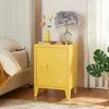 A yellow bedside table with a door and a handle is placed next to a bed. A lamp and a vase with flowers are on top of the table. Produktbild-Vorschau
