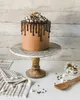 A chocolate cake with marshmallows and chocolate drizzle on a cake stand, with a tea towel and wooden spoon nearby. Anteprima dell’immagine prodotto