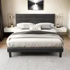 A bedroom scene featuring a dark gray upholstered bed frame with a headboard, dressed with white and gray bedding and decorative pillows. Two white floating nightstands with gold lamps are on either side of the bed. Produktbild-Vorschau