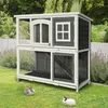 A two-story wooden rabbit hutch is set up on a green lawn with two rabbits in front of it. Voir les images du produit