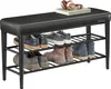 A black upholstered shoe bench with three shelves filled with shoes and boots. A pair of headphones and a book are on top of the bench. Náhled obrázku produktu