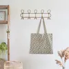 A gold-colored metal coat rack with five hooks is mounted on a white wall, with a patterned tote bag hanging from one of the hooks. A wooden shelf unit with plants and a framed picture is visible to the left, and dried pampas grass is in a vase to the righ Náhled obrázku produktu