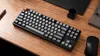 A mechanical keyboard and mouse are placed on a wooden desk, along with a pen and a notebook. Produktbild-Vorschau