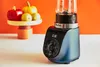 A blue Tefal Blend Up mini-mixer is shown on a white surface with a colorful background. In the background, a pedestal holds red and yellow bell peppers and two green fruits. Anteprima dell’immagine prodotto
