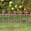 A close-up shot of a decorative metal garden fence with fleur-de-lis finials, set against a blurred background of green foliage and yellow flowers. Produktbild-Vorschau