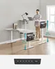 A man stands at a height-adjustable desk, demonstrating its use. The desk is shown in two height configurations: one lower with a book and lamp, and one higher with a laptop and mug. Measurement indicators are present. Produktbild-Vorschau