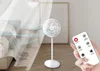 A person is holding a remote control while a white stand fan is operating in a room. Voir les images du produit