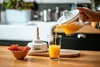 A hand pouring orange juice from a juicer into a glass on a wooden countertop, with a bowl of red currants and a juicer base in the background. Produktbild-Vorschau