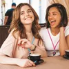 Two young women are sitting at a table outdoors, smiling and laughing while drinking coffee. Produktbild-Vorschau