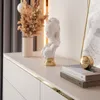 A close-up view of a white bust sculpture with a gold base, placed on a white dresser with gold trim. A vase with dried flowers and a stack of books are also visible. Produktbild-Vorschau