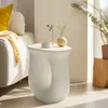 A modern living room scene with a white speckled side table as the focal point, decorated with a small vase and a decorative bowl. A couch with yellow and cream pillows and a draped blanket is visible in the background. Náhled obrázku produktu