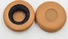 Two brown ear pads for headphones are shown against a white background. One ear pad is shown from the side, revealing its inner black lining and textured fabric. The other ear pad is shown from the top, with a circular mesh opening. Produktbild-Vorschau