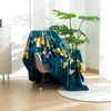 A teal blanket with a gold and white butterfly pattern is draped over a chair. A large green plant is behind the chair. Produktbild-Vorschau