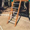 A wooden ladder with metal rungs is set up outdoors on a sandy ground, with trees and shadows in the background. Produktbild-Vorschau