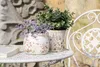 Two decorative pots with artificial plants sit on a white table next to a white chair, with a stone wall in the background. Náhľad obrázku produktu
