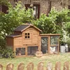 A wooden chicken coop with an attached enclosure is set up in a garden with lush green foliage and a brick building in the background. Two chickens are visible inside the enclosure. Produktbild-Vorschau