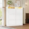 A white shoe cabinet with six flip-down doors and an open shelf with LED lighting on top, decorated with a vase of flowers and framed pictures. Náhľad obrázku produktu