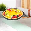 A fruit tart is displayed on a countertop, with a plant in the background and a cutting board and oven mitt to the right. Produktbild-Vorschau