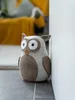 A decorative owl doorstop is placed on the floor next to a white wall, with a blurred background showing a sofa with a yellow throw blanket. Anteprima dell’immagine prodotto