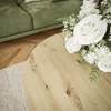 A round wooden coffee table with a bouquet of white roses and baby's breath in front of a green sofa. Produktbild-Vorschau