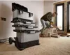 A man is sawing a piece of wood next to a stack of toolboxes. Voir les images du produit